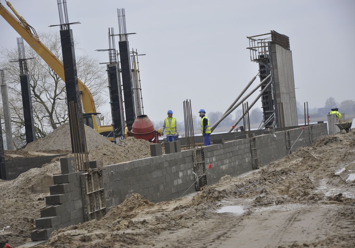 plac budowy, na terenie widać 2 budowniczych obsługujących betoniarkę, w tle widoczny dźwig