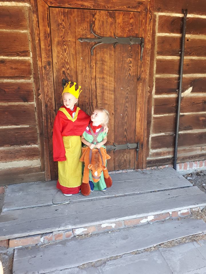 dwójka dzieci przebranych w stroje jedna dziewczynka ma na sobie strój słowiański z pluszową maskotka konikiem, druga czerwono zółty strój  z koroną na głowie. Stoją przed drewnianymi drzwiami do chaty. 