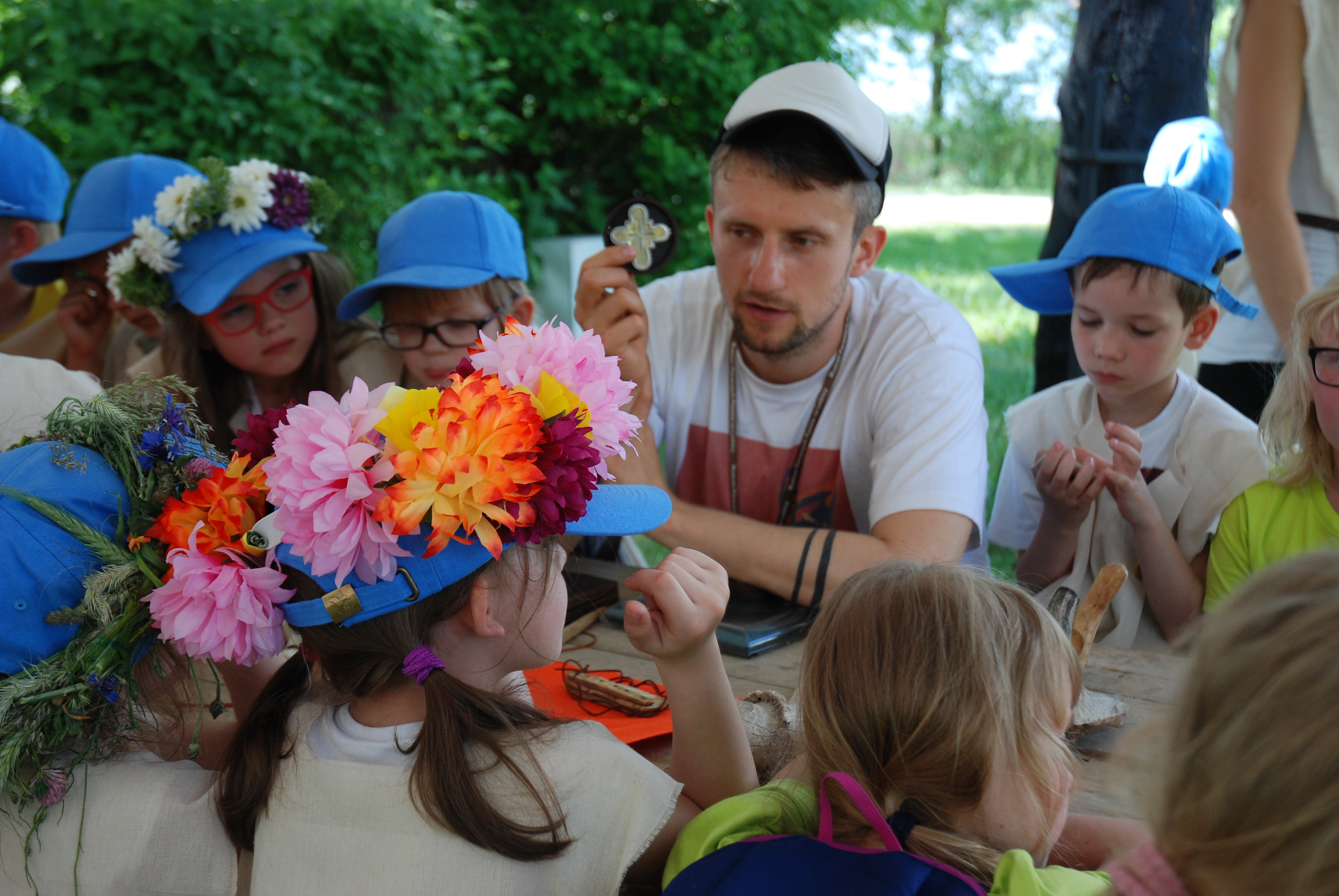 Grupa dzieci siedzących w koło stołu. Na głowach mają równe niebieskie czapki. Kilkoro dzieci ma dodatkowo wianki z kwiatów . Pośrodku nich siedzi edukator, który opowiada trzymając w ręce replikę zabytku stauroteki.  