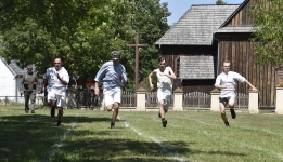 czterech mężczyzn biegnie w tzw. sztafecie. W tle kościół drewniany, zielone drzewa. 
